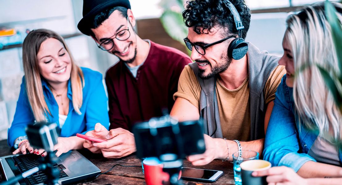  A group of young people are sitting around a table and looking at a laptop and a smartphone, while smiling and laughing, with the intention of creating high-quality and relevant content for Facebook Pro Creators.